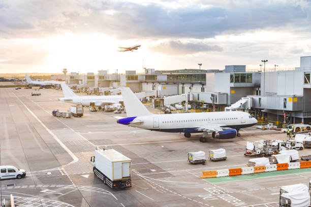 Busy airport view with airplanes and service vehicles at sunset. London airport with aircrafts at gates and taking off, trucks all around and sun setting on background. Travel and industry concepts